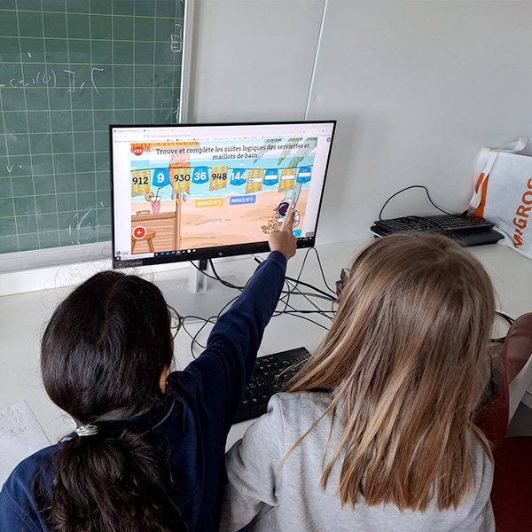 Deux élèves pointant un écran d’ordinateur affichant un exercice de suites logiques en mathématiques.